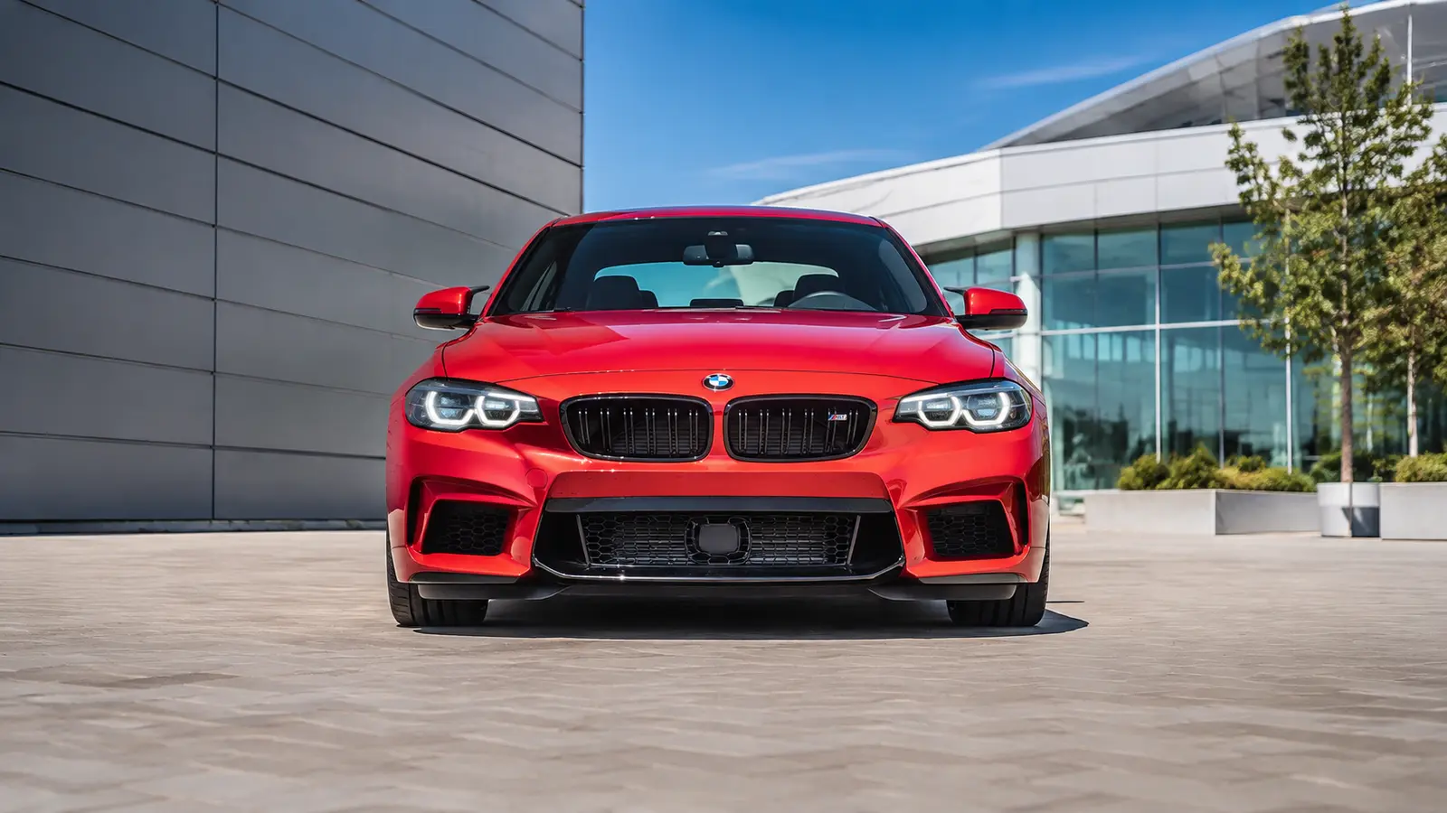 АВТО 1000601 красный BMW M2 строго спереди у современной архитектуры